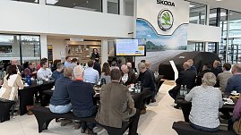 Eine Rednerin steht vor einer Leinwand und präsentiert. Auf dem Bildschirm sind Energiediagramme zu sehen. Gäste hören an langen Frühstückstischen aufmerksam zu. (Foto: Autohaus Peter)