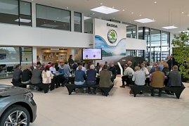 Blick von hinten auf die Gäste beim Unternehmerfrühstück. Vorne ein Bildschirm mit Präsentation, daneben ein schwarzes, abgedecktes Fahrzeug. (Foto: Autohaus Peter)