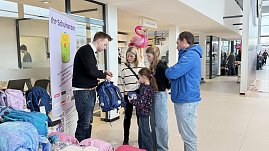 Ein Verkäufer zeigt einer Familie mit zwei Kindern einen blauen Schulranzen. Neben ihnen stehen weitere Ranzen. Im Hintergrund sind Flamingos und Schilder im Autohaus zu sehen. (Foto: Autohaus Peter)