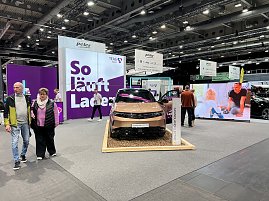 Ein goldfarbener Opel Grandland Electric auf einem Podest mit Holzunterlage, dahinter große lilafarbene Messewand mit Schriftzug „So läuft Laden“. (Foto: Autohaus Peter Gruppe)