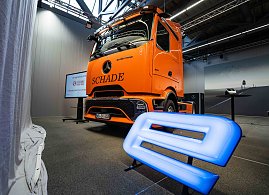 Orangefarbener Mercedes-Benz eActros 600 mit Schade-Logo in einer Halle. Blau leuchtendes E-Symbol im Vordergrund. (Foto: Daimler Truck AG)