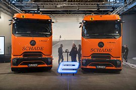 Zwei orangefarbene Mercedes-Benz eActros-Lkw mit Schade-Logo nebeneinander, davor ein blau leuchtendes E-Symbol. Personen im Hintergrund im Gespräch. (Foto: Daimler Truck AG)