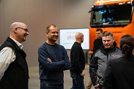 Gruppe von Personen im Gespräch vor einem orangefarbenen Mercedes-Benz eActros mit Schade-Logo. Im Vordergrund Björn Schade im blauen Pullover. (Foto: Daimler Truck AG)
