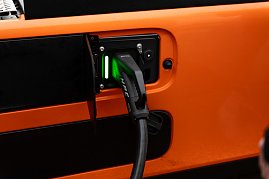 Nahaufnahme eines Ladevorgangs: Ein schwarzes HPC-Ladekabel mit grün leuchtender Anzeige ist an der Seite eines orangefarbenen eActros angeschlossen. (Foto: ex animo MEDIA)