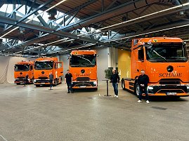 "Vier orangefarbene Mercedes-Benz eActros-Lkw mit Schade-Logo nebeneinander in einer Halle aufgereiht, Personen stehen davor. (Foto: Autohaus Peter)
