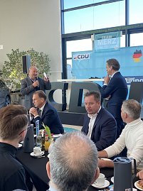 Helmut Peter begrüßt mit Mikrofon die Gäste in der Autowelt Peter. Mehrere Personen sitzen an runden Tischen mit Getränken und Frühstücksgeschirr. Im Hintergrund große CDU-Buchstaben und eine Bühne mit „Team Merz 2025“-Plakat. (Foto: Autohaus Peter)