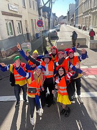 Gruppe von als Narren verkleideten Menschen in orangefarbenen Warnwesten steht auf einer Straße und winkt fröhlich in die Kamera. (Foto: Autohaus Peter GmbH)