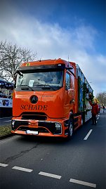 Vorderansicht eines orangefarbenen LKWs mit Aufschrift SCHADE, der den bunt gestalteten Karnevalswagen zieht. (Foto: Autohaus Peter GmbH)