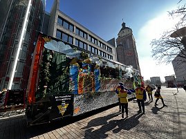Seitliche Ansicht eines bunt dekorierten Karnevalswagens mit Märchenmotiven, gezogen von einem orangefarbenen LKW, im Hintergrund ein historisches Gebäude mit Uhrturm, mehrere als Narren verkleidete Personen laufen daneben. (Foto: Autohaus Peter GmbH)