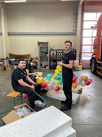 Drei Männer in schwarzer Arbeitskleidung sitzen und stehen in einer Werkstatt, binden bunte Autohaus-Peter-Luftballons zusammen, im Hintergrund ein orangefarbener LKW. (Foto: Autohaus Peter GmbH)
