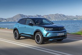 Türkis-schwarzer Opel Mokka Electric fährt an einer Küstenstraße entlang, Meer und Gebirge im Hintergrund, Blick schräg von vorn links. (Foto: Opel Automobile GmbH)
