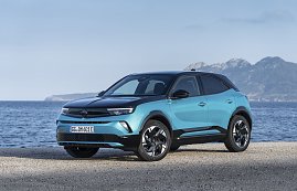 Türkis-schwarzer Opel Mokka Electric steht am Meer, Blick schräg von vorn links, Berge im Hintergrund. (Foto: Opel Automobile GmbH)