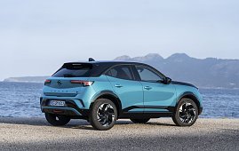 Türkis-schwarzer Opel Mokka Electric steht am Meer, Blick schräg von hinten links, Berge im Hintergrund. (Foto: Opel Automobile GmbH)