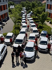 Fuhrparkaufstellung von oben: Reihe von DRK-Fahrzeugen, flankiert von Mitarbeitenden in roten Shirts. Im Vordergrund werden symbolisch Fahrzeugschlüssel und Blumen übergeben. (Foto: Autohaus Peter)