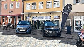 Zwei Mercedes-Transporter  links ein blauer EQV, rechts ein schwarzer Vito  stehen nebeneinander in der Innenstadt von Heiligenstadt. Dazwischen eine schwarze Autohaus-Peter-Flagge, im Hintergrund ein Modegeschäft. (Foto: Autohaus Peter)
