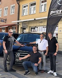 Vier Männer posieren vor einem schwarzen Mercedes Vito auf einer Kopfsteinpflasterstraße in Heiligenstadt. Links und rechts im Hintergrund stehen weitere Fahrzeuge, im Hintergrund sind mehrstöckige Altstadthäuser mit Schaufenstern zu sehen. Neben dem Auto weht eine schwarze Autohaus-Peter-Flagge.  (Foto: Autohaus Peter)