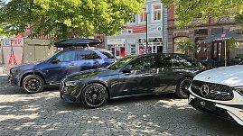 Premiumfahrzeuge in der Innenstadt: Ein blauer Mercedes-Benz SUV mit Dachbox und ein schwarzer Kombi stehen neben weiteren Fahrzeugen auf einem sonnigen Platz in einer Innenstadt mit Cafés und Geschäften. (Foto: Autohaus Peter)