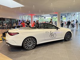 Ein weißes Mercedes-Cabriolet mit der Aufschrift „Shes Mercedes“ steht in einem Autohaus, im Hintergrund sind Gäste und Personal in roten T-Shirts zu sehen. (Foto: Eric Freytag)