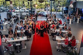 Eine große Gruppe von Frauen sitzt und steht um Tische in einem Autohaus, in der Mitte posiert das „Shes Mercedes“-Team auf dem roten Teppich. (Foto: Eric Freytag)