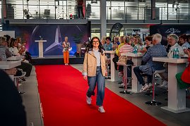 Eine junge Frau mit dunklen, langen Haaren, Brille, beiger Jacke, hellblauem Oberteil, blauen Jeans und weißen Sneakern läuft bei einer Modenschau über einen roten Teppich. (Foto: Eric Freytag)