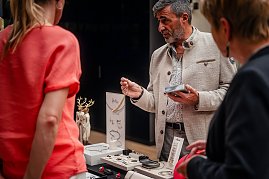 Ein Mann in heller Jacke präsentiert Schmuckstücke an einem Stand, während zwei Frauen interessiert zuhören. Auf dem Tisch liegen Ketten, Armbänder und Ringe. (Foto: Eric Freytag)