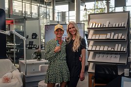 Zwei Frauen lächeln in die Kamera; die linke trägt ein gemustertes Kleid und eine gelbe Kappe, die rechte einen schwarzen Anzug. Im Hintergrund Regale mit Kosmetikprodukten. (Foto: Eric Freytag)