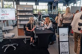 Zwei Frauen sitzen an einem Tisch und unterhalten sich; im Hintergrund steht ein Regal mit Kosmetikprodukten und ein Bildschirm mit der Aufschrift „Shes Mercedes Ladies Night 22. Mai 2025“. Daneben stöbern Besucher an einem Kleiderständer. (Foto: Eric Freytag)