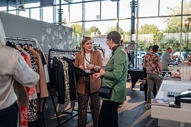 Zwei Frauen unterhalten sich an einem Kleiderständer mit verschiedenen Jacken und Blusen. Die Frau links trägt einen braunen Blazer, die Frau rechts einen grünen Blazer. (Foto: Eric Freytag)