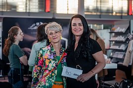 Geschäftsführerin Christina Dost im schwarzen Hosenanzug und Moderatorin Mandy Kossira im bunten, geblümten Blazer lächeln gemeinsam in die Kamera, Christina Dost hält eine Karte mit „Shes Mercedes“-Logo. (Foto: Eric Freytag)