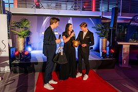 Drei Personen stehen auf rotem Teppich auf einer Bühne, Christina Dost spricht ins Mikrofon, während ein Mann ein Los aus einem goldenen Hut zieht. (Foto: Eric Freytag)