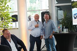 Afrikanischer Abend im Autohaus Peter: Helmut Peter und Abenteuer-Guide Hannu begrüßen die Gäste lachend, während Helmut Peter das Mikrofon hält. Links im Bild lacht ein weiterer Teilnehmer, rechts ist ein Stehtisch mit südafrikanischer Dekoration und ein Bildschirm mit einem Safari-Motiv zu sehen. (Foto: Autohaus Peter)