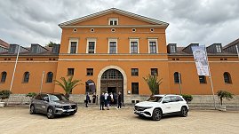 Vorderansicht des historischen Gebäudes mit orangefarbener Fassade. Zwei Mercedes-Benz Fahrzeuge stehen auf dem Platz davor, während sich einige Besucher am Eingang unterhalten. Rechts ist ein Banner mit Werbung für ein kulturelles Event zu sehen. (Foto: Autohaus Peter)