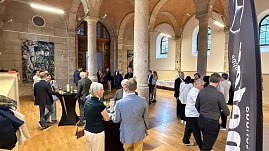 Innenaufnahme eines historischen Saals mit hohen Steinsäulen und Holzboden. Gäste stehen an Stehtischen, unterhalten sich und genießen Getränke. Im Hintergrund ist ein Buffet aufgebaut. (Foto: Autohaus Peter)