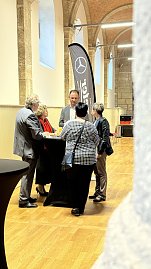 Eine kleine Gruppe von fünf Personen unterhält sich im historischen Saal an einem schwarzen Stehtisch. Im Hintergrund sind hohe Steinsäulen, Wände aus Naturstein und ein großes Mercedes-Benz Werbefähnchen zu sehen. (Foto: Autohaus Peter)