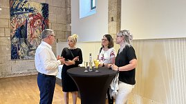 Vier Personen stehen und unterhalten sich an einem schwarzen Stehtisch in einem historischen Saal mit Holzboden und Steinwänden. Auf dem Tisch stehen Gläser mit Sekt und Orangensaft. Im Hintergrund hängt ein farbenfrohes Gemälde an der Wand. (Foto: Autohaus Peter)