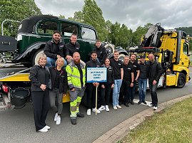 Gruppenfoto eines Teams des Autohaus Peter beim Festumzug in Sondershausen. Die Mitarbeitenden stehen vor einem klassischen Abschleppwagen des ADAC mit einem Oldtimer auf der Ladefläche. Alle tragen schwarze Autohaus-Peter-Bekleidung, einige lachen in die Kamera. Im Vordergrund wird ein Schild mit der Aufschrift „Autohaus Peter GmbH“ gehalten. Im Hintergrund Bäume und bewölkter Himmel. (Foto: Autohaus Peter)