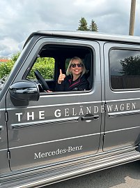 Eine lächelnde Frau mit Sonnenbrille sitzt am Steuer eines grauen Mercedes-Benz G-Klasse Geländewagens, lehnt sich aus dem offenen Fenster und zeigt den Daumen hoch. Auf der Fahrzeugseite ist die Aufschrift „THE GELÄNDEWAGEN“ und „Mercedes-Benz“ gut sichtbar. (Foto: Autohaus Peter)