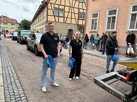 Zwei Mitarbeiter der Peter Gruppe stehen lächelnd auf der Straße während des Festumzugs in Sondershausen. Hinter ihnen fahren Fahrzeuge der Firma, am Straßenrand stehen Zuschauer. (Foto: Autohaus Peter)