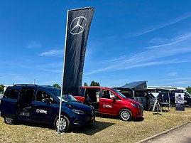 Mehrere Mercedes-Benz Fahrzeuge des Autohauses Peter, darunter ein blauer Van, ein roter Van und ein silberner Campingbus mit geöffnetem Aufstelldach, stehen auf einer Ausstellungsfläche beim Anhalt 2025 Leichtathletik-Meeting. Vor den Fahrzeugen weht eine schwarze Fahne mit Mercedes-Stern, rechts stehen Infotafeln. Der Himmel ist klar und blau. (Foto: Autohaus Peter )