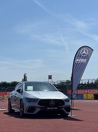 „Ein silberner Mercedes-AMG steht auf der Laufbahn des Anhalt 2025 Leichtathletik-Meetings. Rechts neben dem Fahrzeug weht eine schwarze Fahne des Autohauses Peter im Wind. Im Hintergrund sind Tribünen und Werbebanner zu sehen, während der Himmel klar und sonnig ist. (Foto: Autohaus Peter )