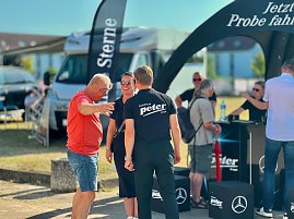 Menschen unterhalten sich gut gelaunt am Stand des Autohauses Peter beim Anhalt 2025 Leichtathletik-Meeting. Im Vordergrund stehen zwei Personen in schwarzer Autohaus-Peter-Kleidung und ein Mann im roten T-Shirt. Im Hintergrund sind weitere Besucher, ein Wohnmobil und schwarze Promotion-Zelte mit Mercedes-Benz-Logo zu sehen. (Foto: Autohaus Peter )