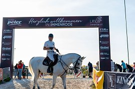 Ein Reiter in weißem T-Shirt und schwarzem Helm sitzt auf einem weißen Pferd unter einem Begrüßungsbogensorenn mit der Aufschrift Herzlich willkommen in Immenrode. Der Bogen ist mit Logos verschiedener Spo versehen. Im Hintergrund sind mehrere Personen und ein sonniger Himmel zu sehen. (Foto: Autohaus Peter)