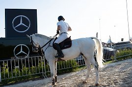 „Ein Reiter in weißer Turnierkleidung sitzt auf einem weißen Pferd vor einem Mercedes-Benz Werbebanner mit zwei großen Logos. Der Reitplatz ist von Absperrgittern und grünen Sträuchern umgeben, im Hintergrund stehen Fahrzeuge und Eventzelte. (Foto: Autohaus Peter)