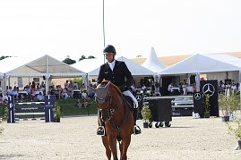 Ein Reiter in schwarzer Turnierjacke und Helm reitet auf einem braunen Pferd über den Sandplatz. Im Hintergrund sitzen Zuschauer unter weißen Pagodenzelten, daneben sind Hindernisse und Mercedes-Benz Werbetafeln zu sehen. (Foto: Autohaus Peter)