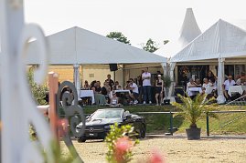 „Zuschauer sitzen unter weißen Eventzelten an Tischen und verfolgen ein Springreitturnier. Vor den Zelten steht ein schwarzes Mercedes-Benz Cabrio, dahinter blühende Pflanzen und ein sonniger Himmel. (Foto: Autohaus Peter)