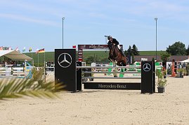 Ein Reiter in schwarzer Reitkleidung und Helm springt mit einem braunen Pferd über ein Hindernis mit Mercedes-Benz-Logo. Im Hintergrund sind Zuschauer unter einem weißen Zelt, mehrere Fahnen und eine grüne Landschaft zu sehen. (Foto: Autohaus Peter)