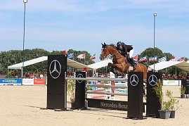 Ein Reiter springt mit einem braunen Pferd über ein Mercedes-Benz-Hindernis auf dem Sandplatz beim Reitturnier in Immenrode. Im Hintergrund sitzen Zuschauer unter weißen Tribünendächern, darüber wehen zahlreiche Länderflaggen. (Foto: Autohaus Peter)