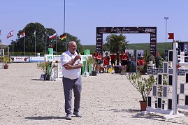 Helmut Peter spricht mit einem Mikrofon auf dem Springplatz beim Reitturnier in Immenrode. Hinter ihm stehen Helfer in roten Shirts vor dem Begrüßungsbogen und wehenden Fahnen. (Foto: Autohaus Peter)