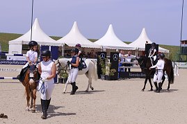 Mehrere Kinder reiten auf Ponys beim Reitturnier in Immenrode, begleitet von Erwachsenen in Reitkleidung. Im Hintergrund stehen weiße Pagodenzelte und ein Hindernis mit Mercedes-Benz-Werbung. (Foto: Autohaus Peter)
