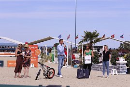 Siegerehrung beim Reitturnier in Immenrode: Auf dem Sandplatz stehen fünf Erwachsene, einer spricht ins Mikrofon. Vor ihnen liegen Preise wie ein Kinderfahrrad und Geschenktaschen, im Hintergrund sitzen Zuschauer auf den Tribünen unter wehenden Fahnen. (Foto: Autohaus Peter)
